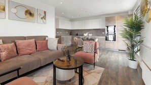 A modern living room with a sofa, armchairs, and a coffee table. A kitchen with a dining area is visible in the background. - The Lasgidi - One Bed (Barking)