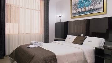 A neatly made bed with a white and brown blanket, a flat-screen TV on a stand, and a painting of trees on the wall.