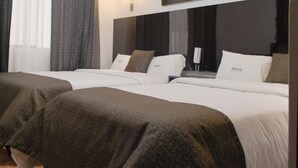 Hotellihuone, jossa on kaksi sänkyä, seinällä puuta kuvaava taulu, yöpöytä lampun kanssa ja ikkuna, jossa on verhot.