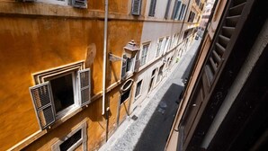 A narrow street with buildings on both sides, featuring multiple windows with shutters and a cobblestone pavement.