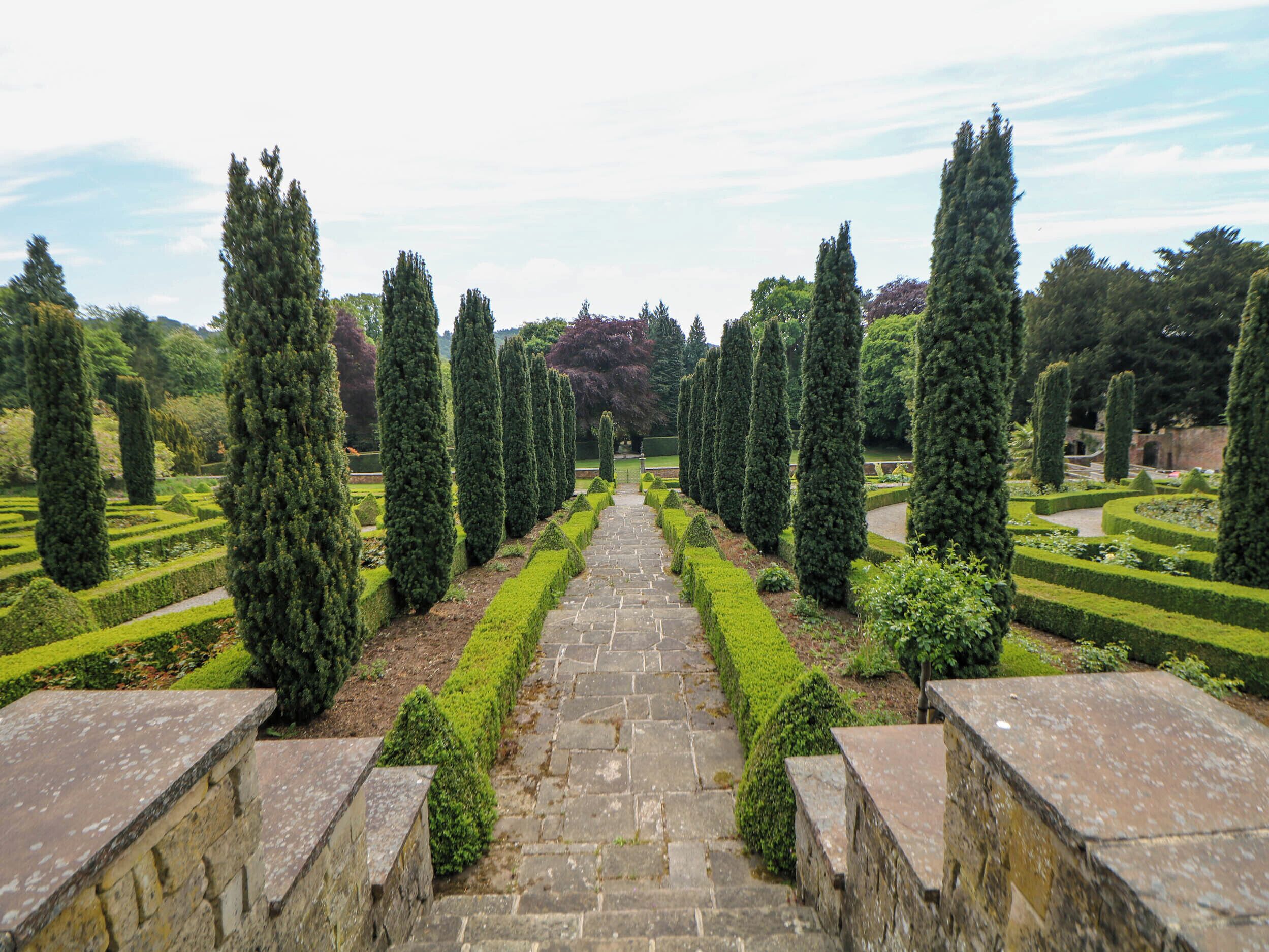 A formal garden with a stone pathway leading through neatly trimmed hedges and tall trees.