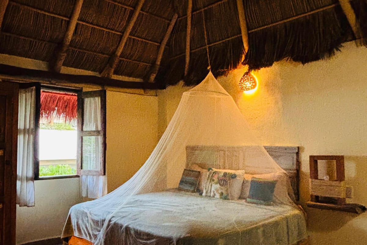 A room with a bed, a window with curtains, a bedside table, and a thatched roof.