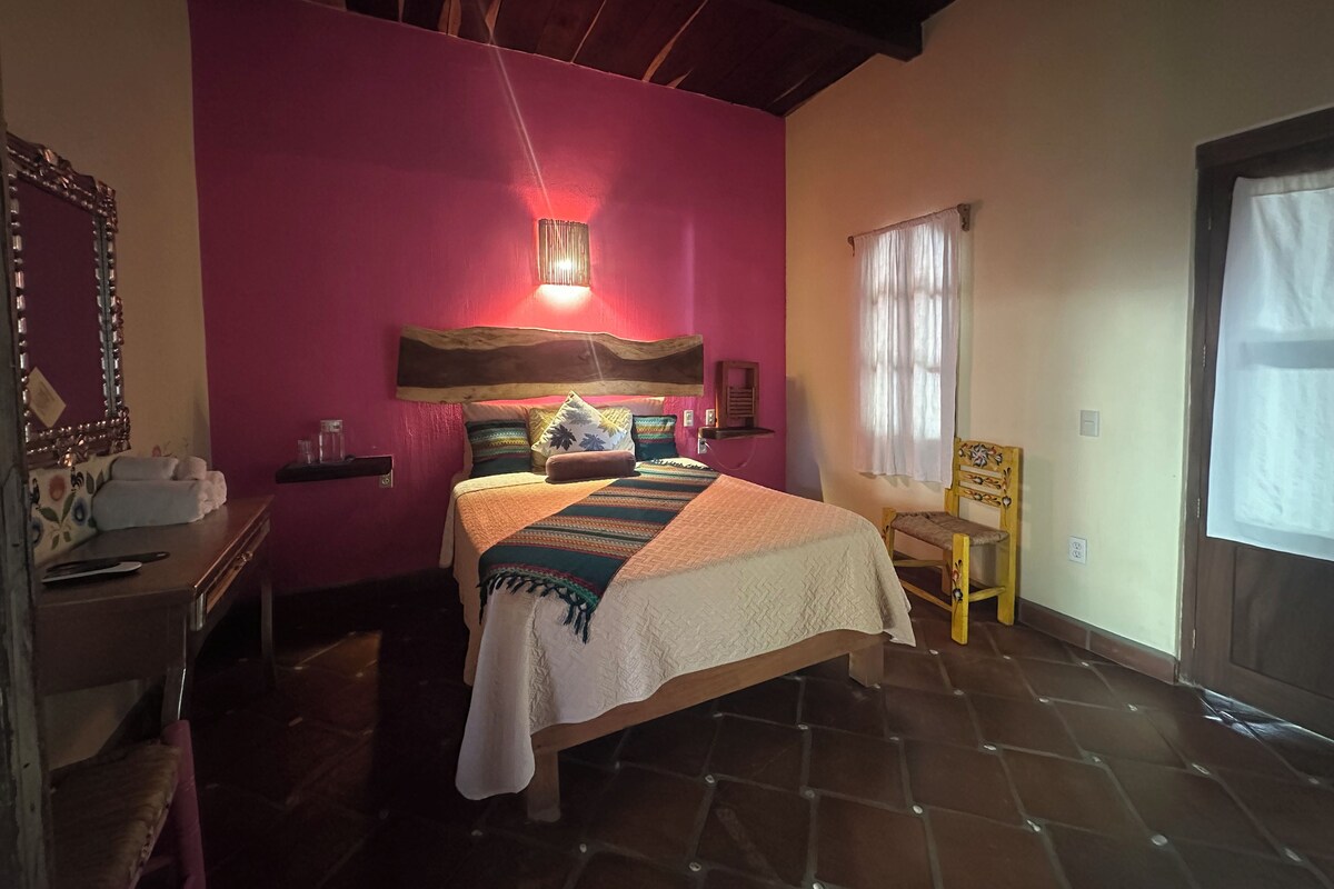 A bedroom with a bed, a nightstand, a chair, a window, and a mirror.