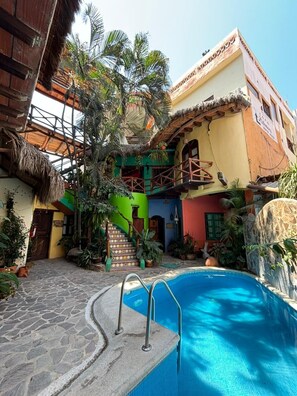 Un área de piscina con un sendero de piedra, una estructura de madera y un edificio colorido con una escalera.