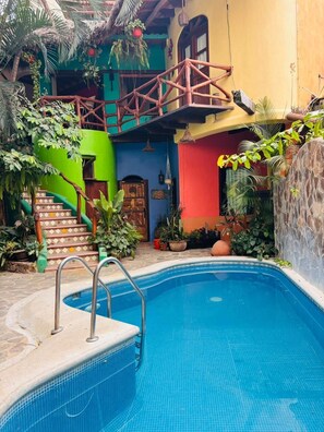 Una piscina con un puente de madera, rodeada de edificios coloridos y exuberante vegetación.