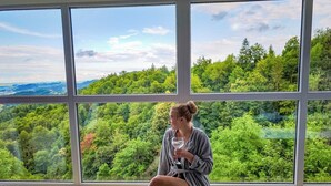 Une personne assise près d’une grande fenêtre, d’où l’on peut admirer un paysage boisé.