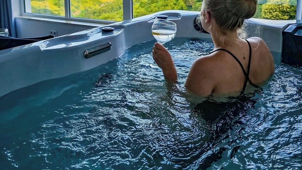 Une personne se détend dans un bain à remous, avec vue sur un paysage verdoyant grâce à de grandes fenêtres.