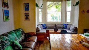 A cozy living room with a wooden table, a sofa, and a window with a view of greenery. - Green Elephant Backpackers (Cape Town)