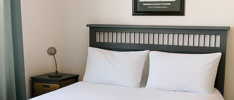 Una cama bien hecha con ropa de cama blanca y azul, un cabecero oscuro y un cuadro en la pared.