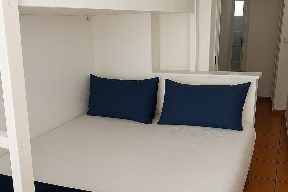 Una cama con ropa de cama blanca y dos almohadas azul oscuro en una habitación con paredes blancas y suelo de baldosas.