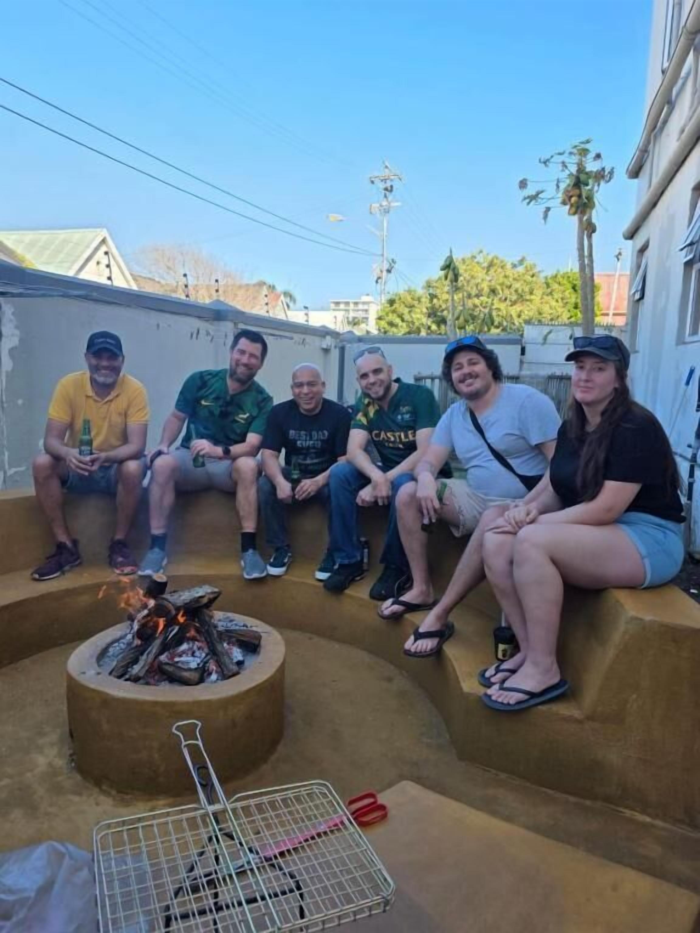Un grupo de personas sentadas alrededor de un brasero en un entorno exterior, con una parrilla de metal y unas tijeras en el suelo.