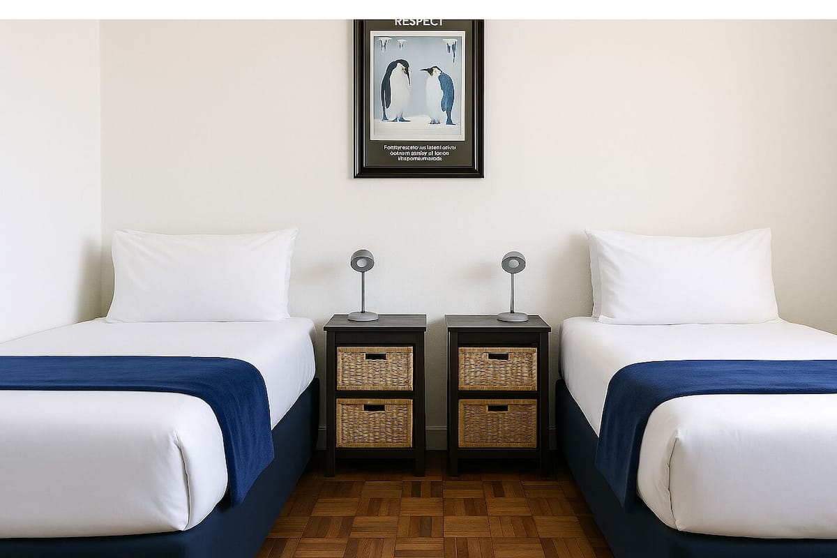 Habitación de hotel con dos camas, una mesita de noche con lámpara y un cuadro enmarcado en la pared.