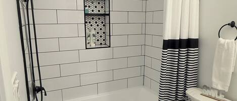 A bathroom with a white tiled shower, a black and white striped shower curtain, a white toilet, and a small window above the shower.