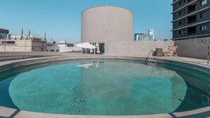 En swimmingpool med en klar blå vandoverflade, omgivet af en betonflisebelagt terrasse og en metalstige.