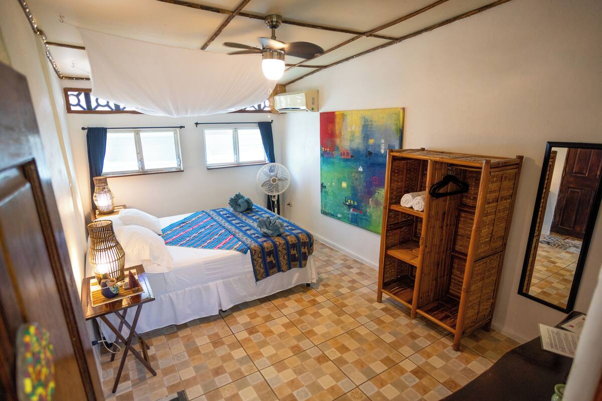 A bedroom with a bed, a fan, a wardrobe, and a painting on the wall.