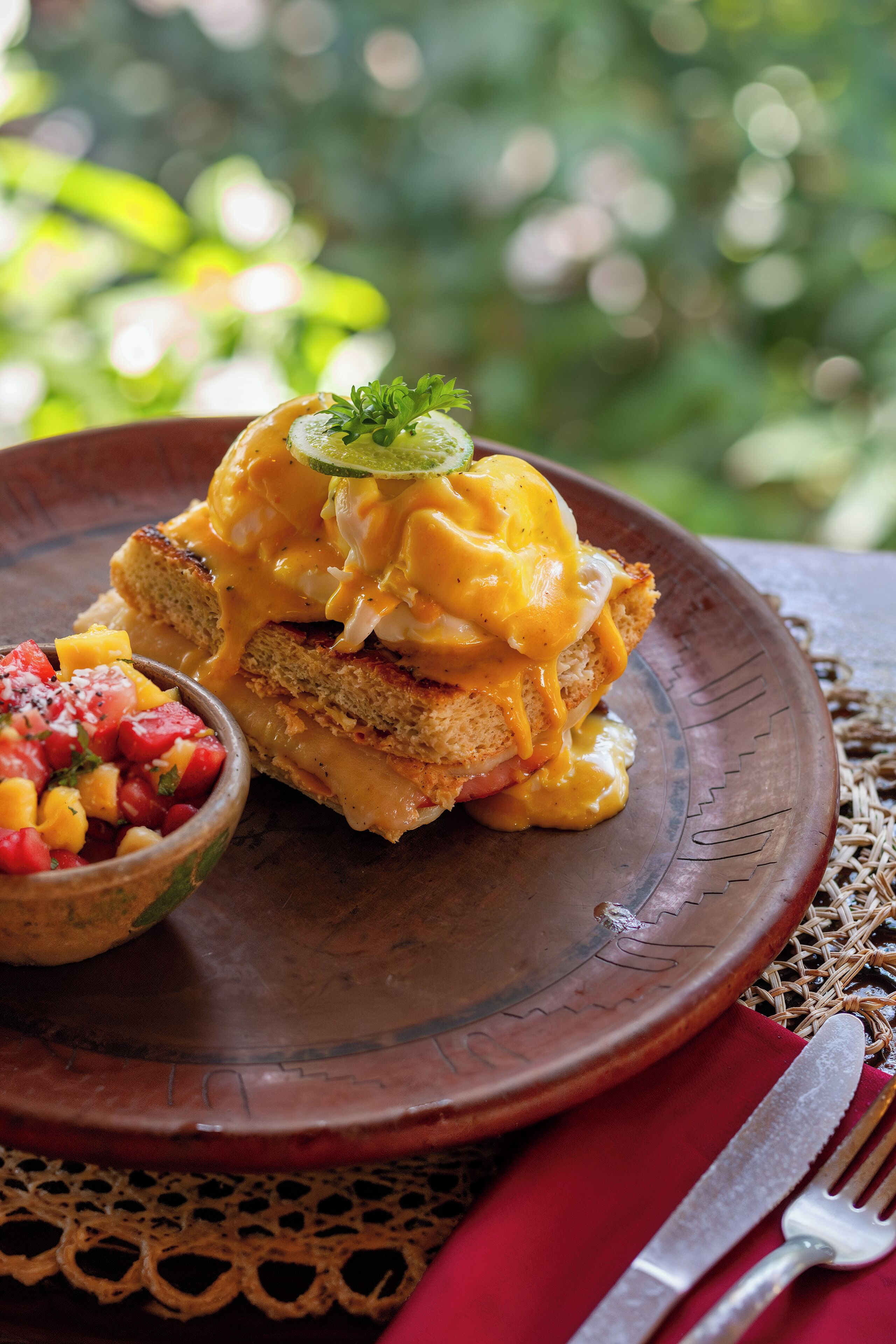 Prato de pequeno-almoço com ovos Benedict, salada de frutas e um guardanapo vermelho com talheres sobre uma mesa rústica.
