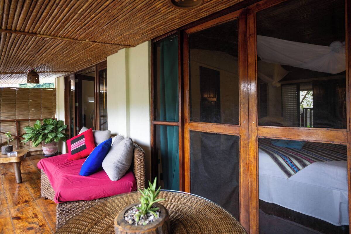 A room with a woven ceiling, a sofa with cushions, a small table with a plant, and a large window with a curtain.