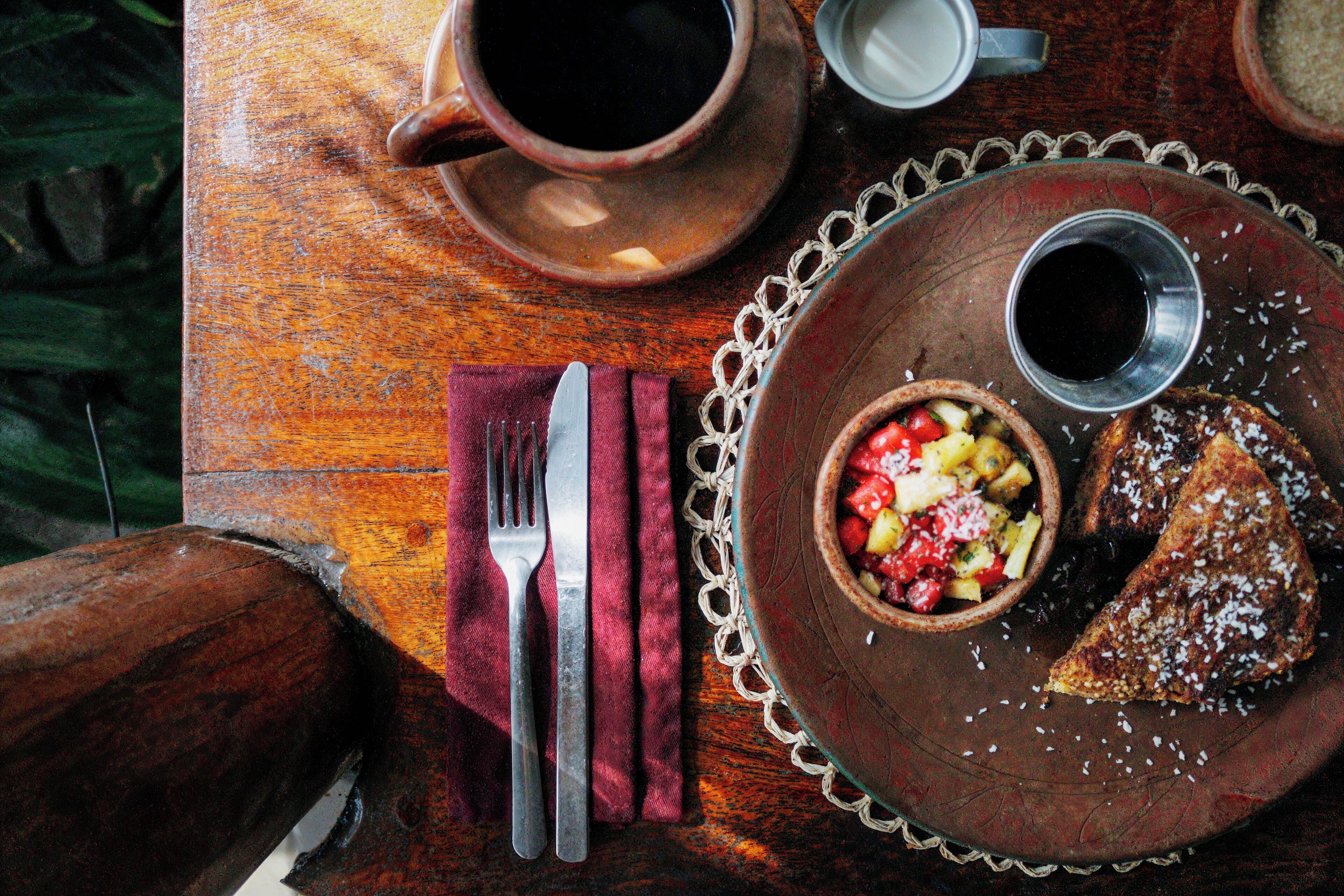 Une table en bois, dans un décor rustique, avec une assiette contenant de la nourriture, une tasse, ainsi qu’une fourchette et un couteau.