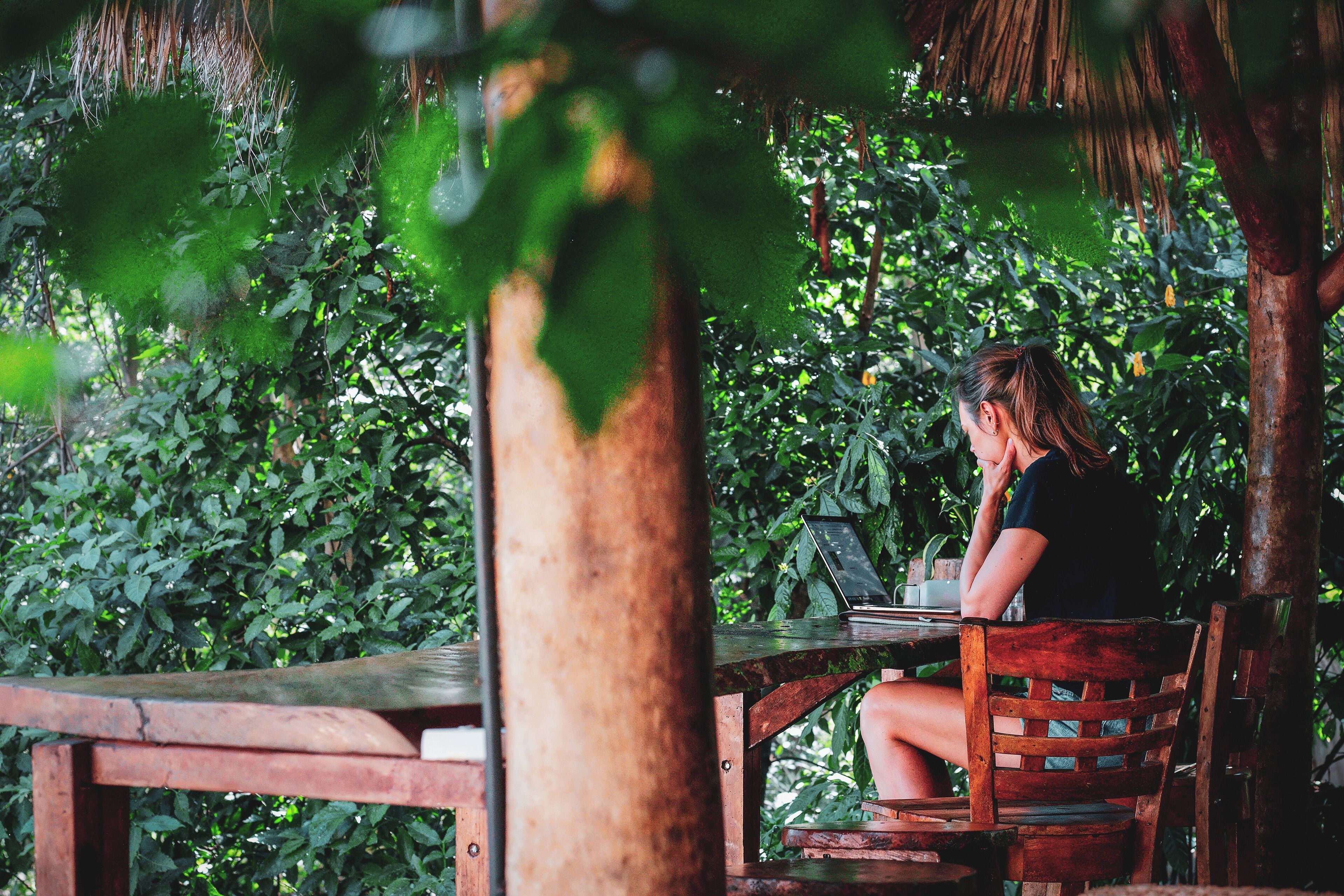 Une personne est assise à une table en bois, un ordinateur portable devant elle, et elle est entourée de verdure.