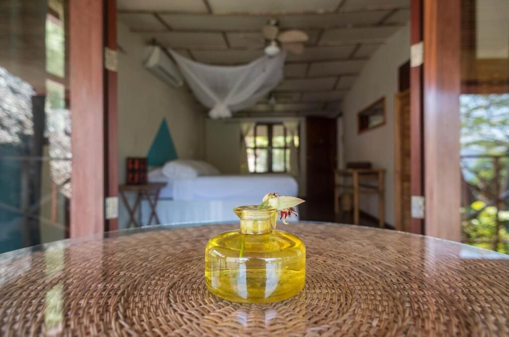 A room with a bed, a hanging white cloth, and a bottle of oil on a woven table.