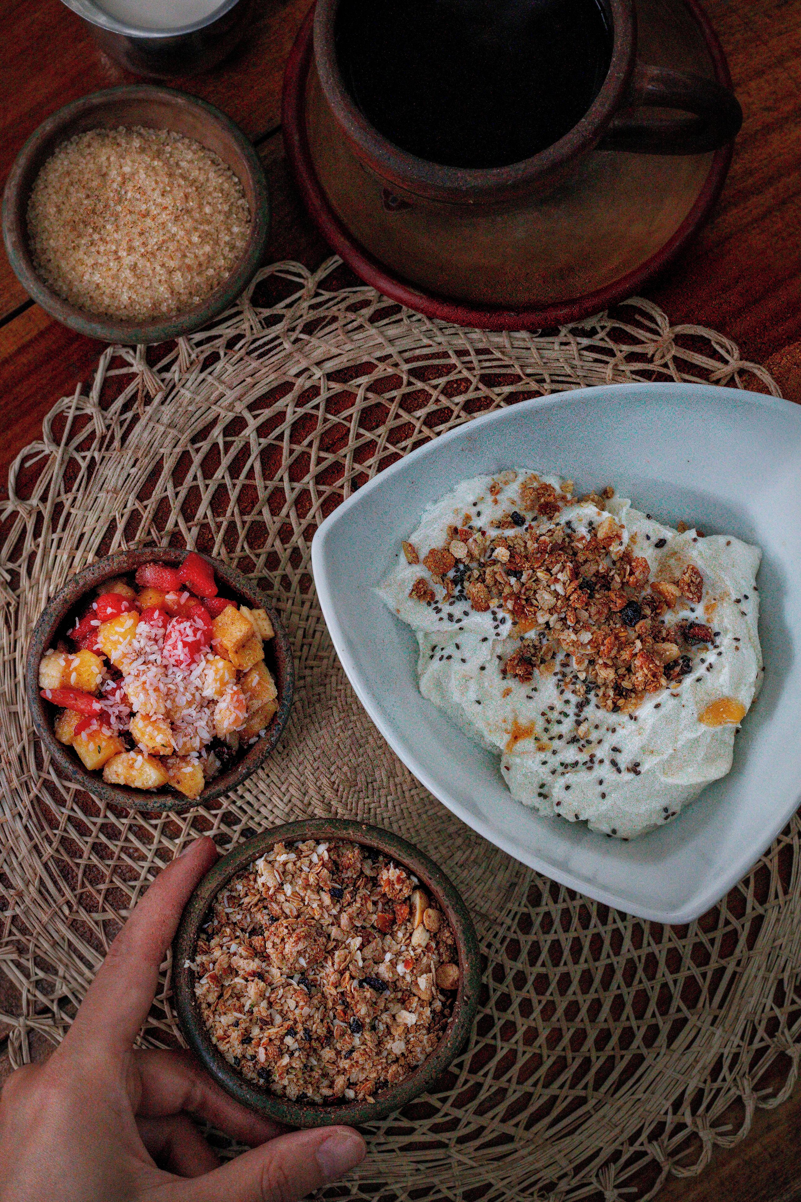 A table with a woven placemat, a bowl of granola, a bowl of yogurt with fruit and granola, and a rustic teapot.