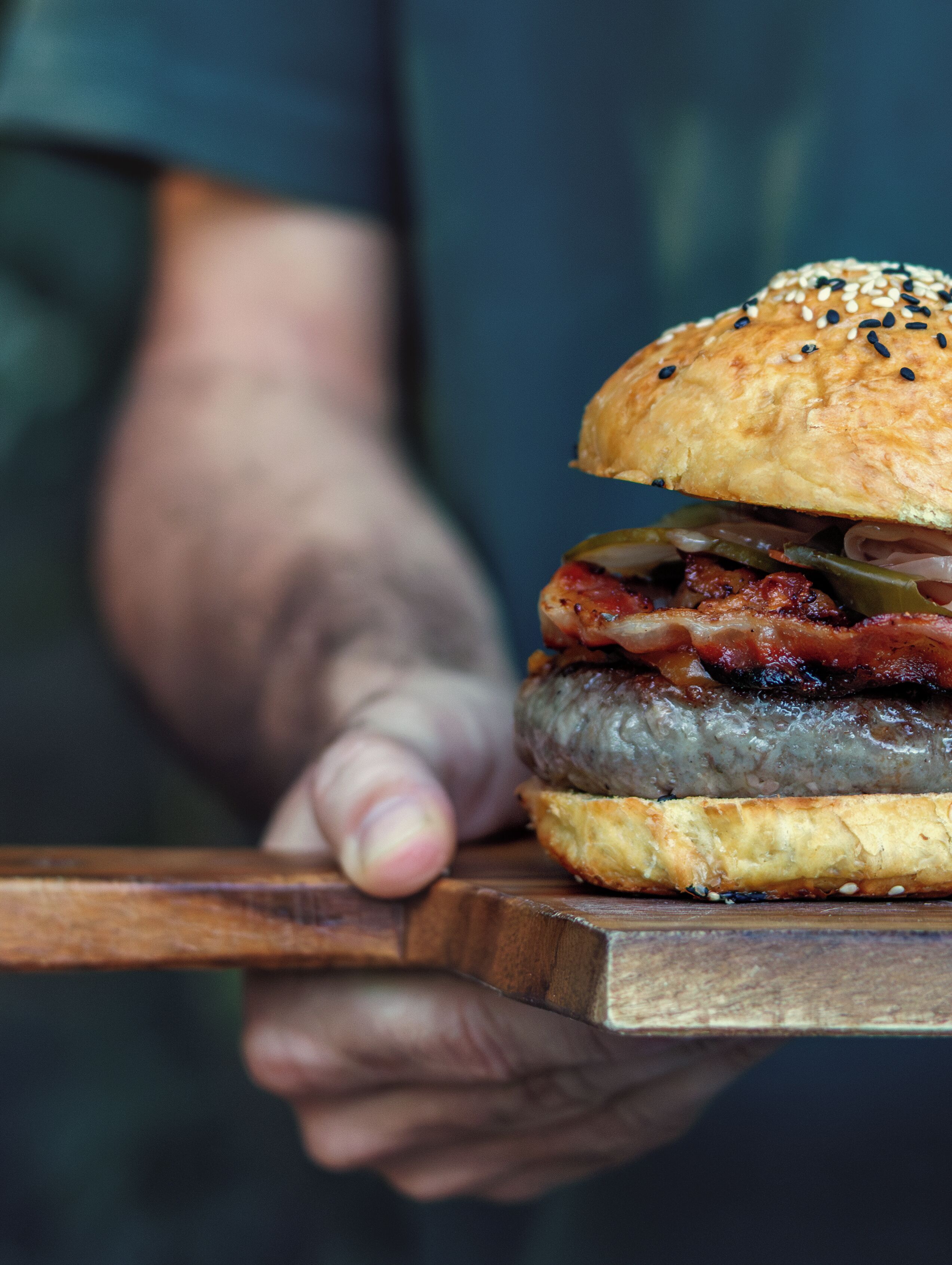 A person holding a burger with a sesame seed bun and bacon.