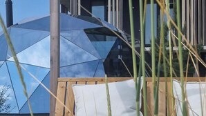 Une terrasse en bois avec un espace salon agrémenté de coussins blancs, une structure en forme de dôme de verre et une pergola en bois en arrière-plan.