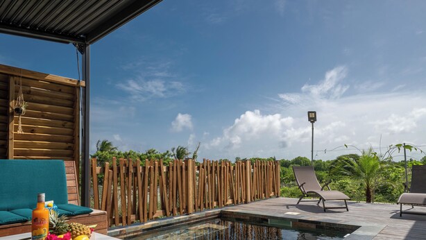A pool area with clear water, a wooden deck, lounge chairs, and a table with fruits and a bottle.