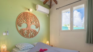 A bedroom with a bed, a wall-mounted air conditioner, a window with a view, and a decorative tree wall art piece.