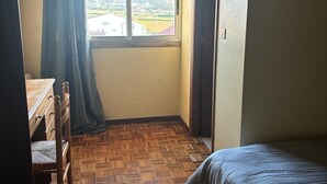 Una habitación con piso de madera, una cama, un escritorio con silla, un ventanal con cortinas y una puerta.