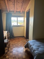 Una habitación con piso de madera, una cama, un escritorio con silla, un ventanal con cortinas y una puerta.