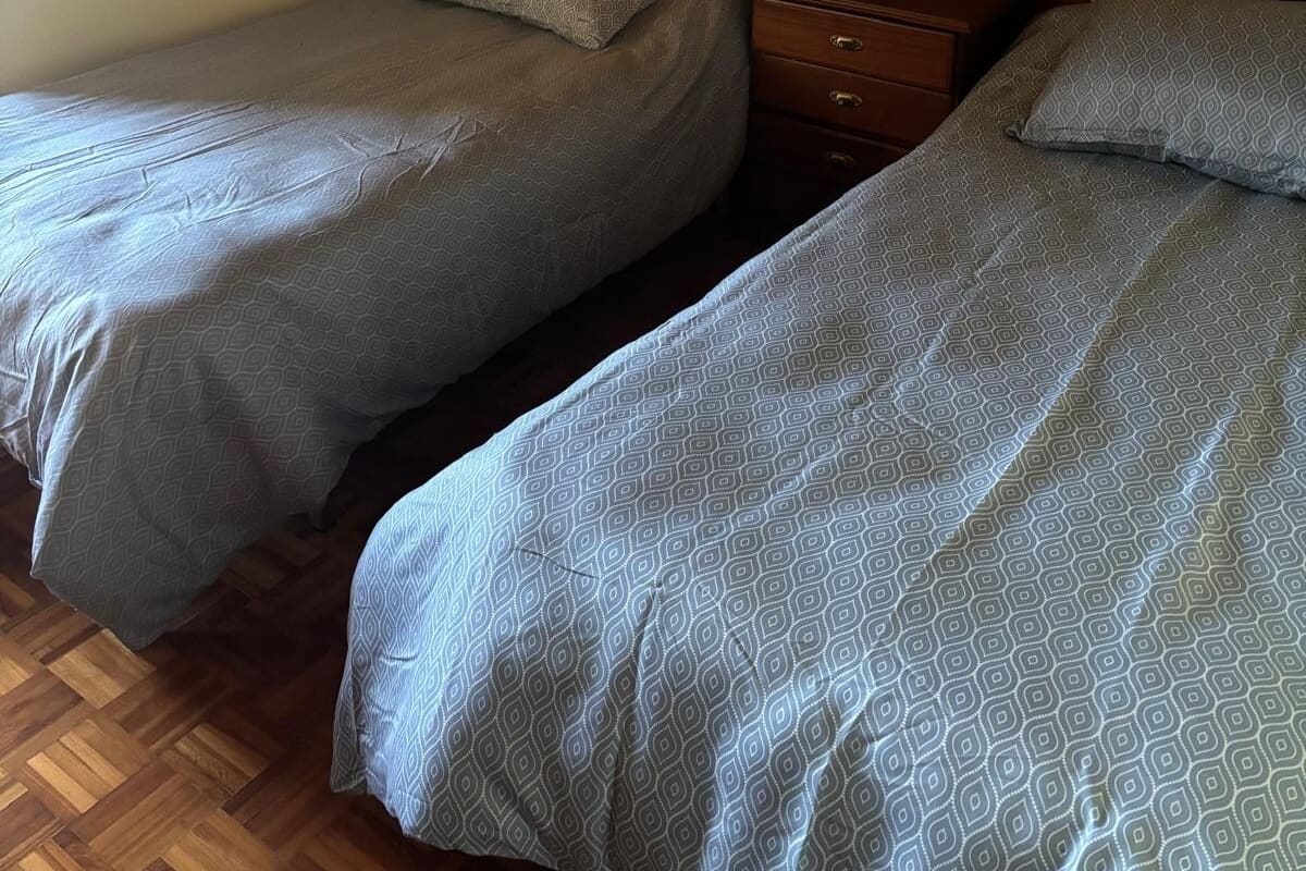 A room with two single beds, each with a patterned duvet and pillows, a wooden nightstand with a lamp, and a wall-mounted electrical outlet.