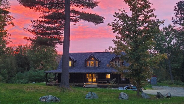 A two-story log cabin with a porch and a small attached shed, surrounded by trees and rocks.