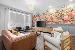 Un salon moderne doté d’une grande fenêtre, d’un téléviseur à écran plat, d’une fresque murale à motifs floraux et de canapés en cuir.