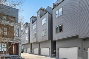 A row of modern multi-story residential buildings with grey siding, multiple garage doors, and a clear sky.