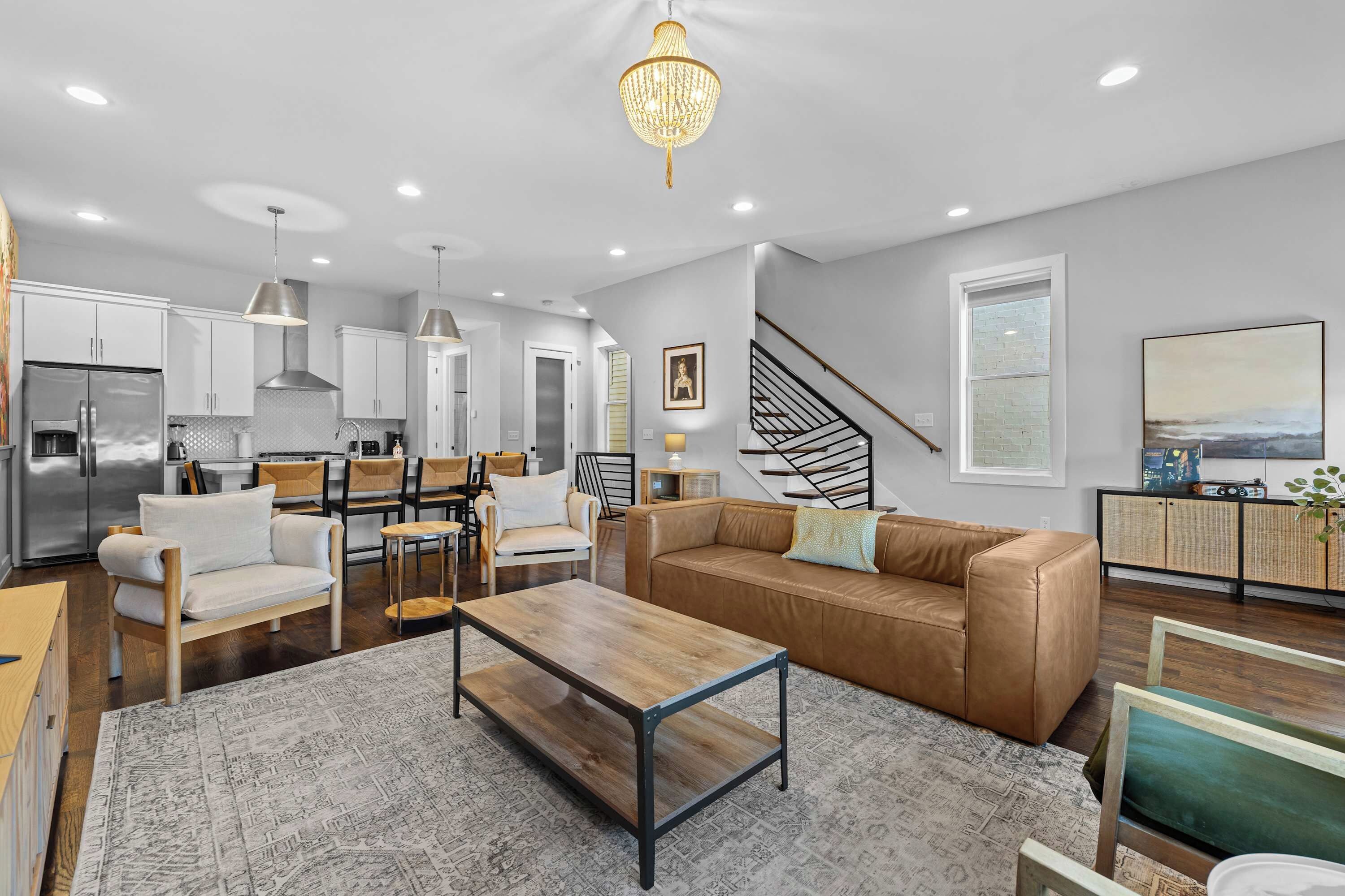 A modern kitchen with stainless steel appliances, a dining area with a wooden table and chairs, a leather sofa, and a staircase leading to another level.