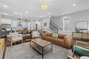 A modern kitchen with stainless steel appliances, a dining area with a wooden table and chairs, a leather sofa, and a staircase leading to another level.