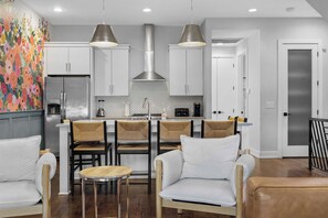Une cuisine moderne avec des armoires blanches, un réfrigérateur en acier inoxydable et une cuisinière à gaz. Deux luminaires suspendus sont placés au-dessus de l’îlot central, et un coin repas comprend une table ronde et des chaises.