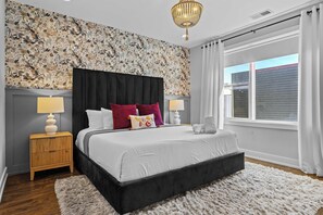 A bedroom with a large bed, two bedside tables, a chandelier, a window with curtains, and a floral-patterned wall.