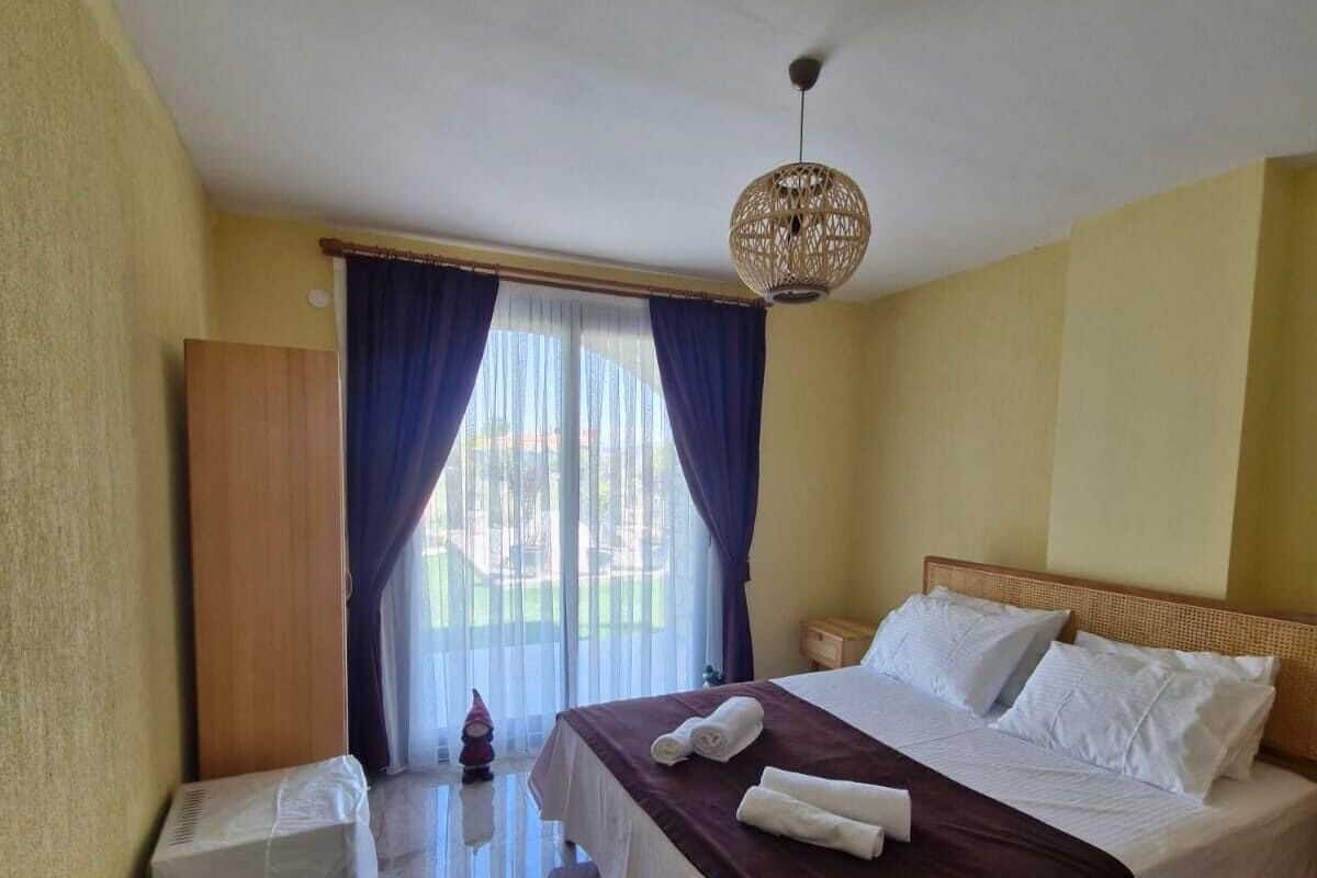 A hotel room with a bed, a small trash bin, a decorative gnome, and a large window with curtains.