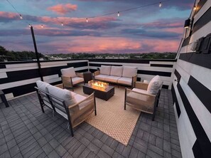 Een terras op het dak met een vuurkuil, tuinmeubilair en lichtjes, gelegen tegen een zonsondergang.