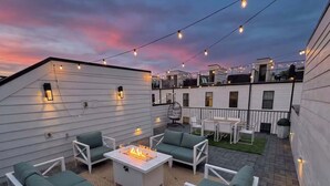 Una terrazza sul tetto con mobili da esterno, un braciere, luci a catena e vista sulla città al crepuscolo.
