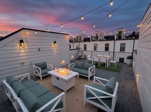 Une terrasse sur le toit, aménagée avec des meubles d’extérieur, un foyer, des guirlandes lumineuses et offrant une vue sur le paysage urbain au crépuscule.