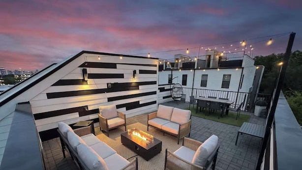 Una terrazza sul tetto con mobili da esterno, un braciere e luci a stringa sullo sfondo di un cielo al tramonto.