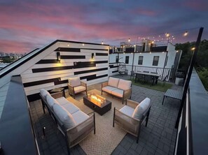 A rooftop patio with outdoor furniture, a fire pit, and string lights against a sunset sky.