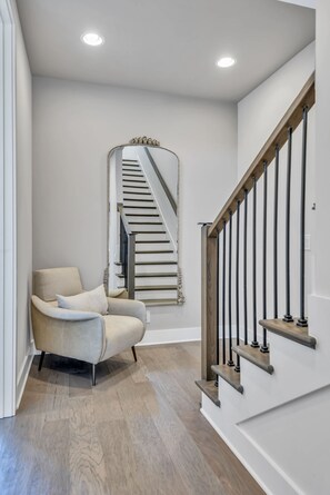 A modern staircase with wooden steps and black balusters, a beige armchair with a cushion, and a mirror with a decorative frame. - Luxe Oasis ~2mi to DT• Skyline • Rooftop • Firepit (Nashville)
