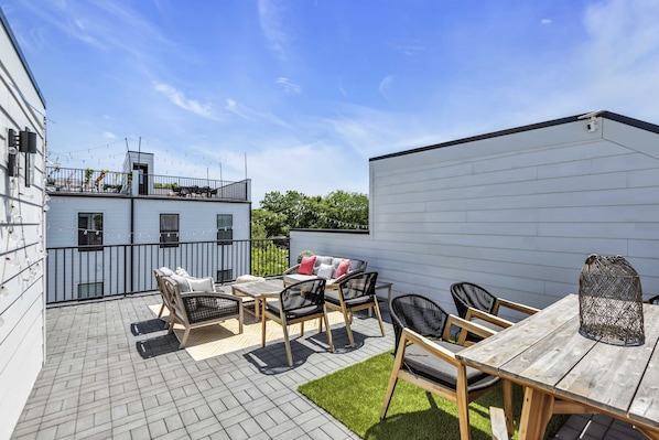 An outdoor patio area with a wooden table and chairs, a sofa with cushions, and a small garden.