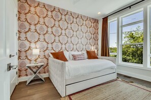 A bedroom with a white bed, brown pillows, a patterned wallpaper, and a large window with a view of trees.