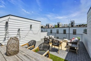 A rooftop patio with outdoor furniture, a wooden table, and a white building in the background.