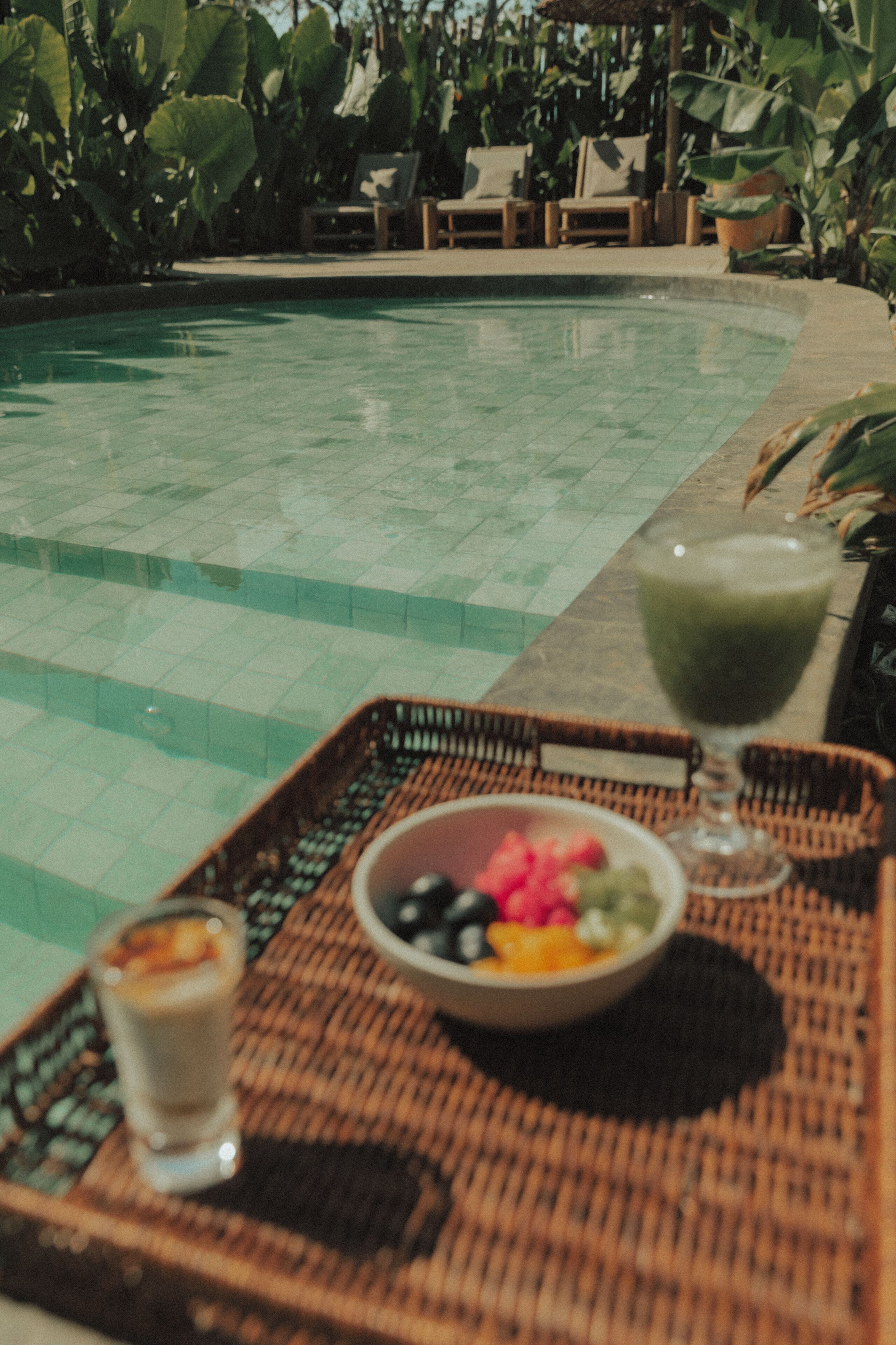 A poolside setting with a wicker tray holding a bowl of fruit and a drink, and a glass of a beverage.